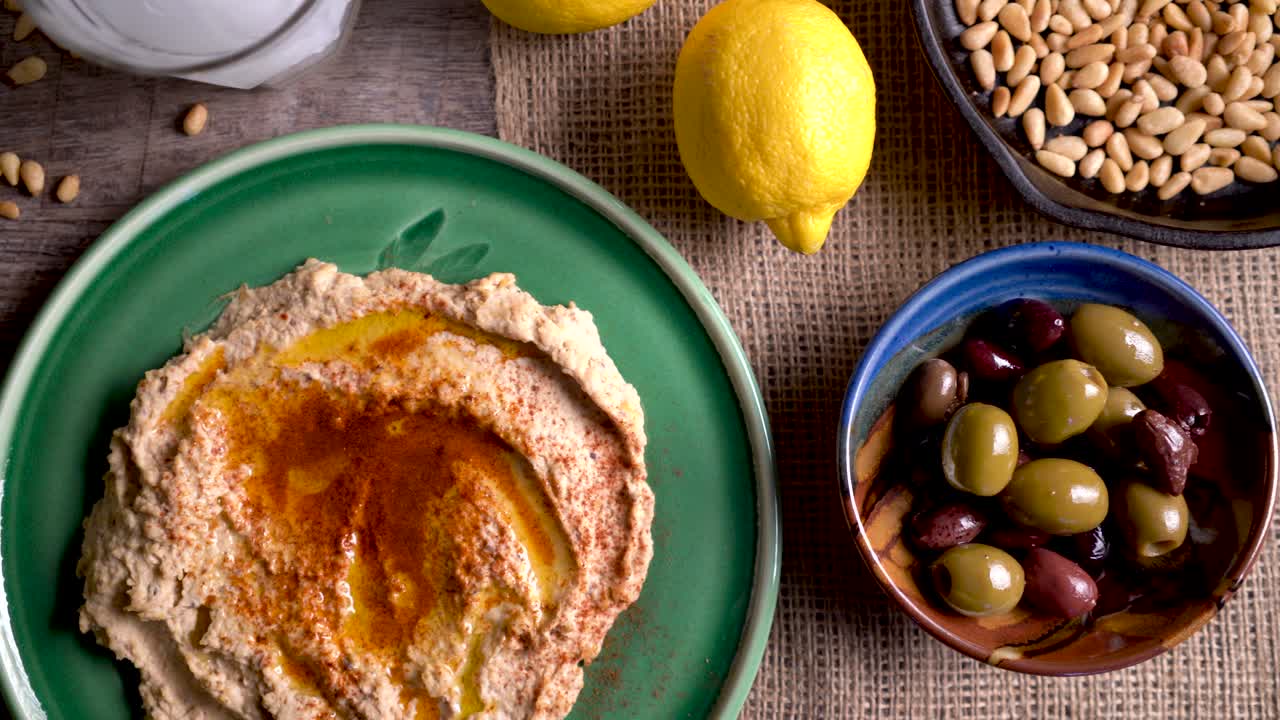 Scorrendo verso destra, viene mostrato un piatto di hummus con feta, olive, un bicchiere di ouzo, limoni e pinoli su un tavolo di legno rustico