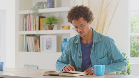 man relaxing at home sitting at table reading book with hot drink