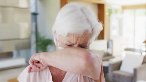 senior caucasian woman wearing face mask sneezing on her elbow at home