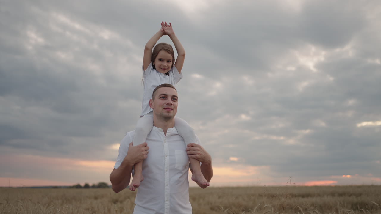 Papà porta sulle spalle la sua amata figlioletta sana sotto il sole. Al rallentatore, la bambina cammina con il padre nel campo e, libera e felice, agita le mani verso l'alto. Passeggiata nel campo.