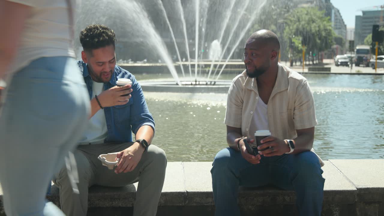 Two men sitting on fountain ledge, left man offering cup, right man taking, holding phone, chatting