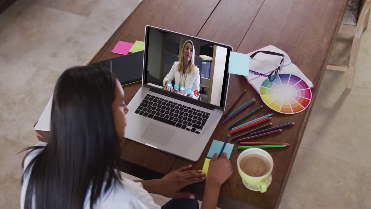 Mujer caucásica usando una laptop en videollamada con una colega, tomando notas