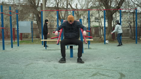 a coach adjusts himself on outdoor workout equipment, while two boys play football in the background, one boy in a gray sweater kicks the ball away from the boy in black