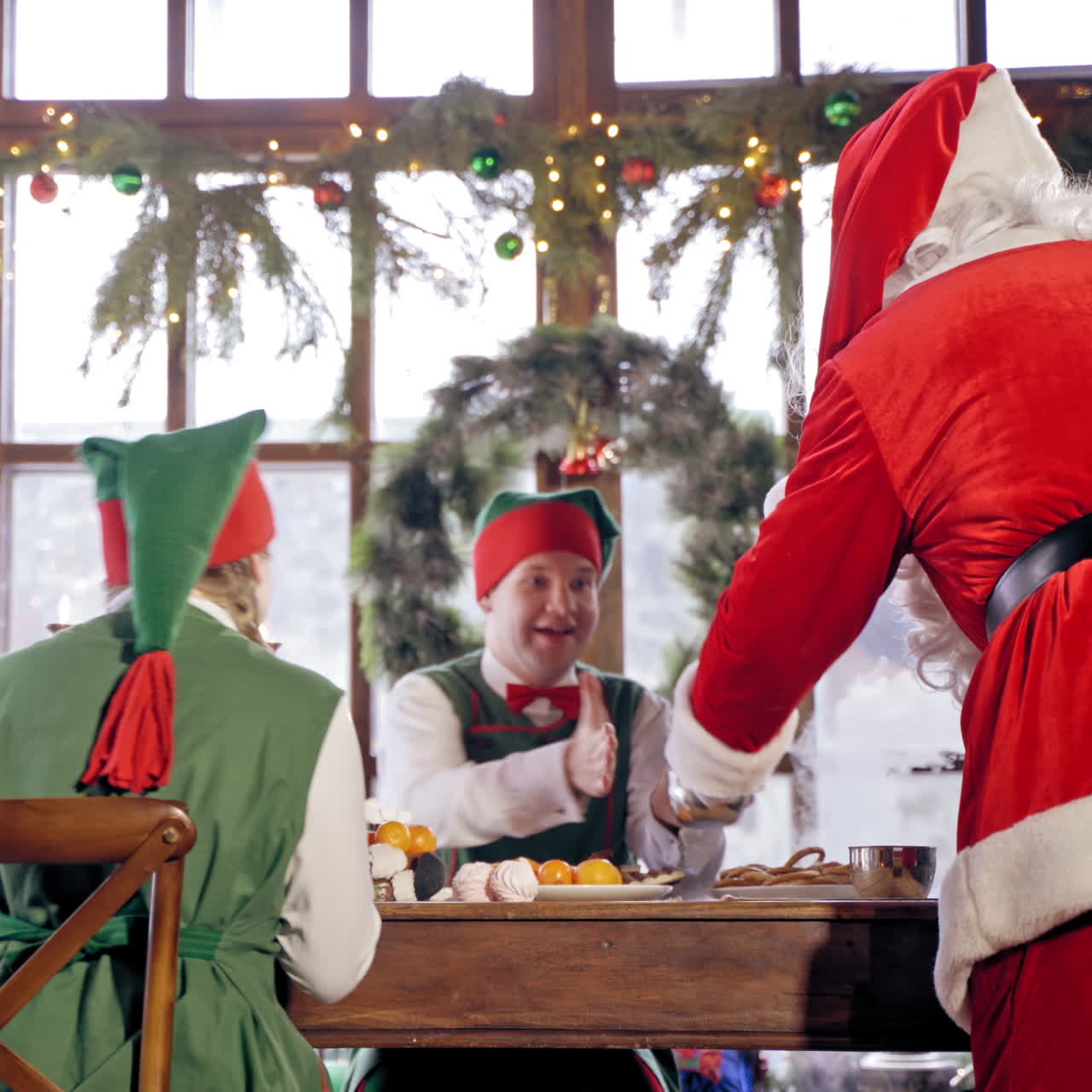 Santa Claus is pouring hot tea to the cups in specially decorated room. Happy elves are looking at the process. New Year and Christmas concept. Feast is coming.