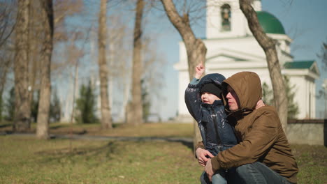 father squatting and holding his son on his leg, the child, dressed in a shiny black jacket, is pointing excitedly at something in the distance, with houses and trees visible