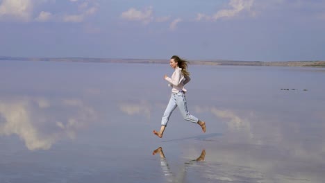 Feliz-Mujer-De-Cabello-Largo-En-Casual-Está-Corriendo-En-La-Arena-Poco-Profunda-Del-Lago-Con-Reflejo-En-La-Superficie-Del-Espejo