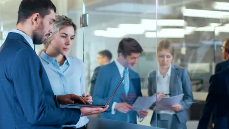 un hombre de negocios con una computadora portátil habla con una mujer de negocios en el vidrio de la oficina, discutiendo temas relacionados con el negocio. gente de negocios ocupada caminando por el pasillo del edificio corporativo.
