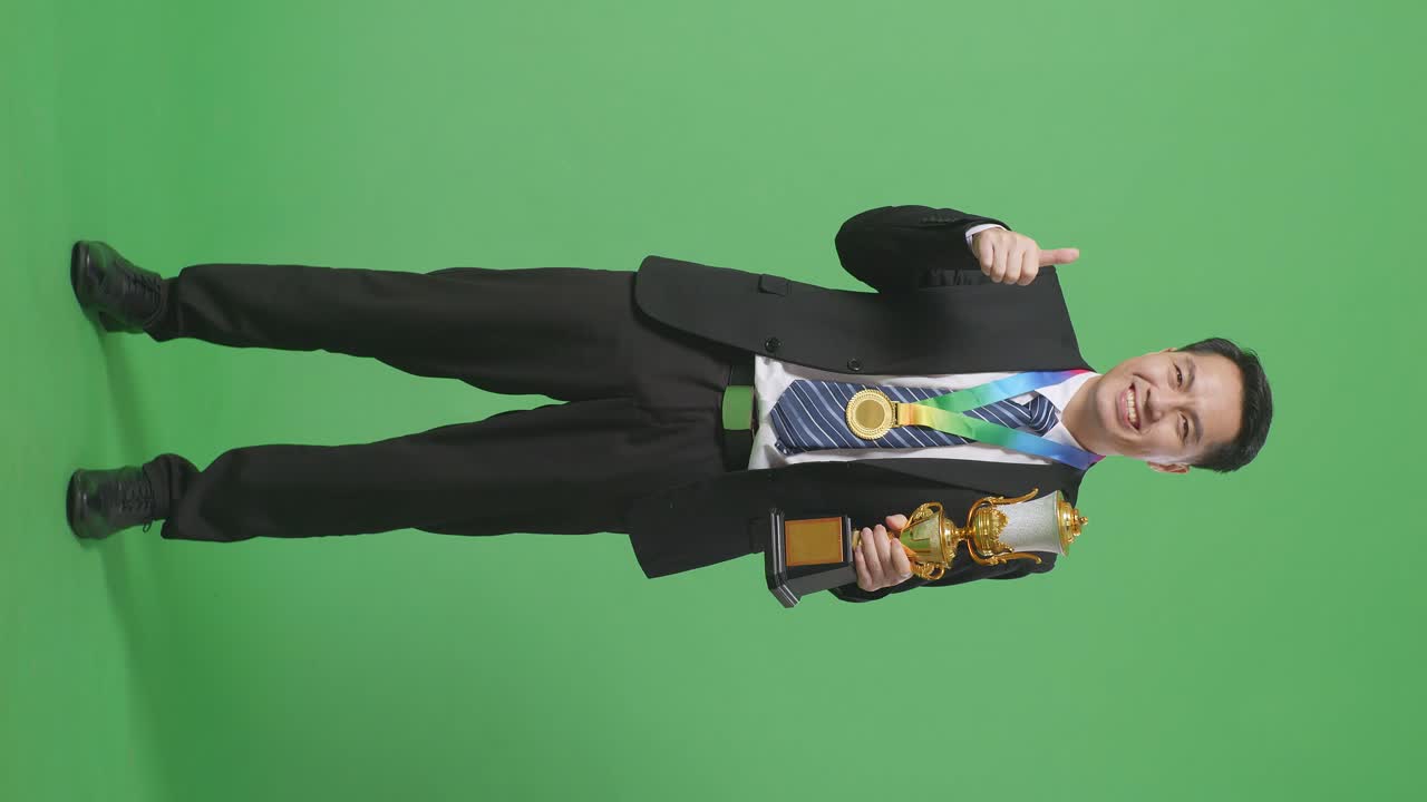 슈트와 타이를 입은 아시아 사업가, 금메달과 트로피를 들고, 엄지손가락을 올리고, 카메라에 미소를 지으며, 스튜디오의 녹색 스크린 배경에서 첫 번째 우승자입니다.
