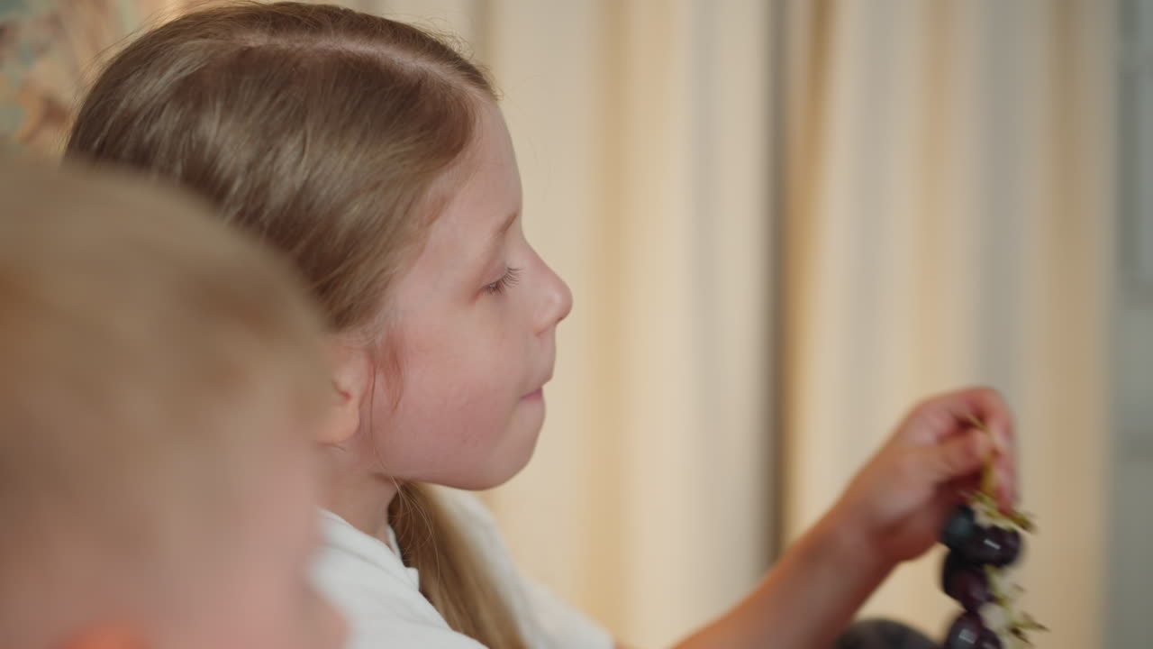 cute girl eating fresh black berries directly from branch while little boy sits beside her indoors cozy atmosphere with natural daylight soft beige curtain background