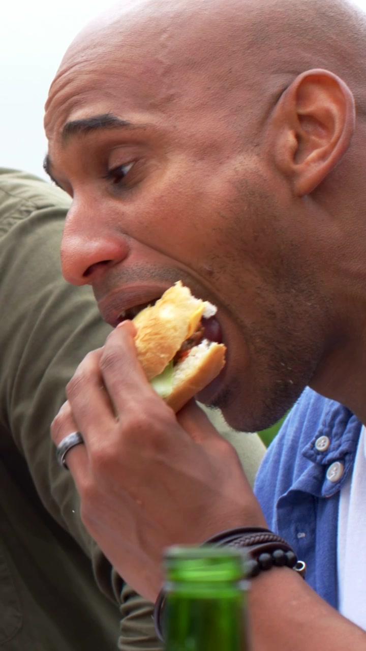 Vertical video: Talking, bald man in blue shirt leaning in, picking sandwich and eating outdoors