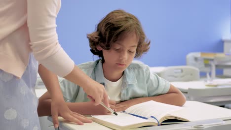 boy reading textbook in class. a girl helps him and explains the exercises to do. medium shot. child sitting at his desk.