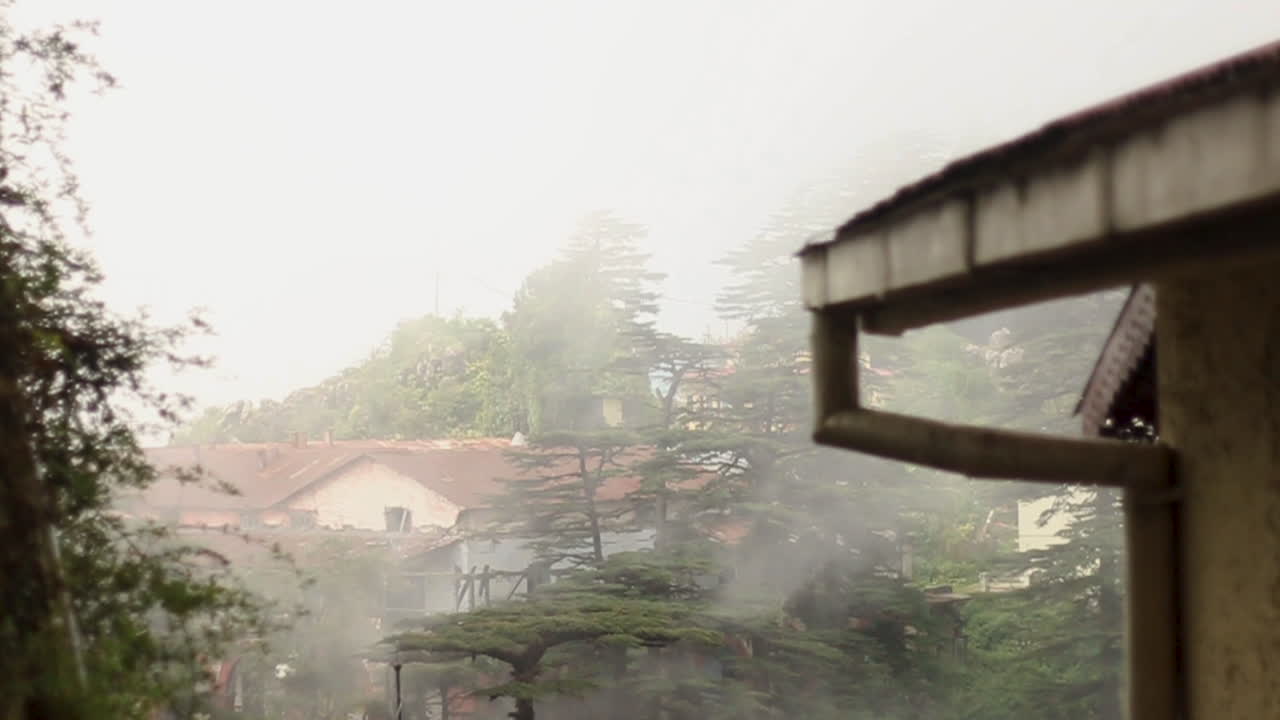 Shift focus onto dense fog movement against a scenic background of houses and tall trees in Mussoorie, slow motion handheld shot.