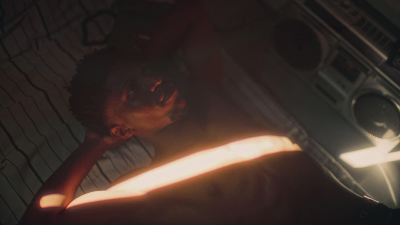 Shirtless Black Man Resting on Bed Illuminated by Beam of Sunlight