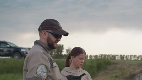male and female field workers dressed in matching uniforms interact while standing on grassy roadside, man holds device and looks upward, woman gestures thoughtfully, with black vehicle parked