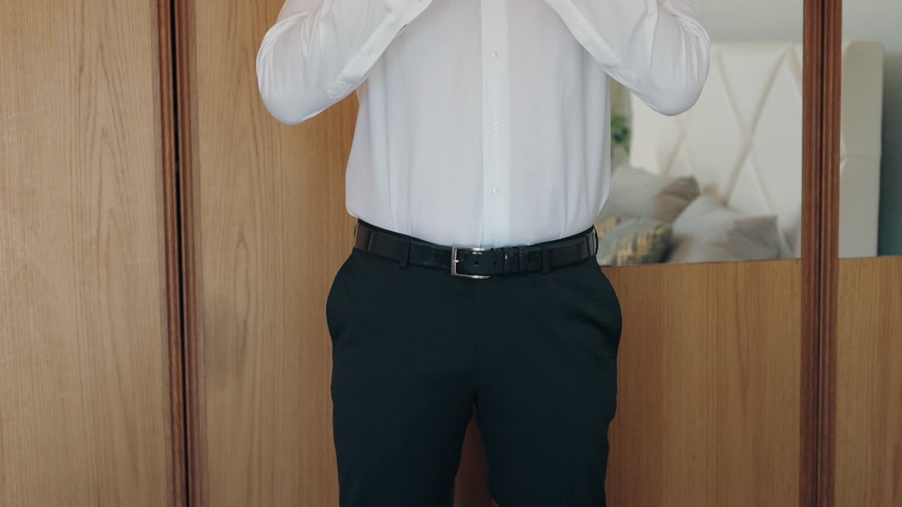 Close up of man buttoning white shirt with black trousers and belt in front of wardrobe