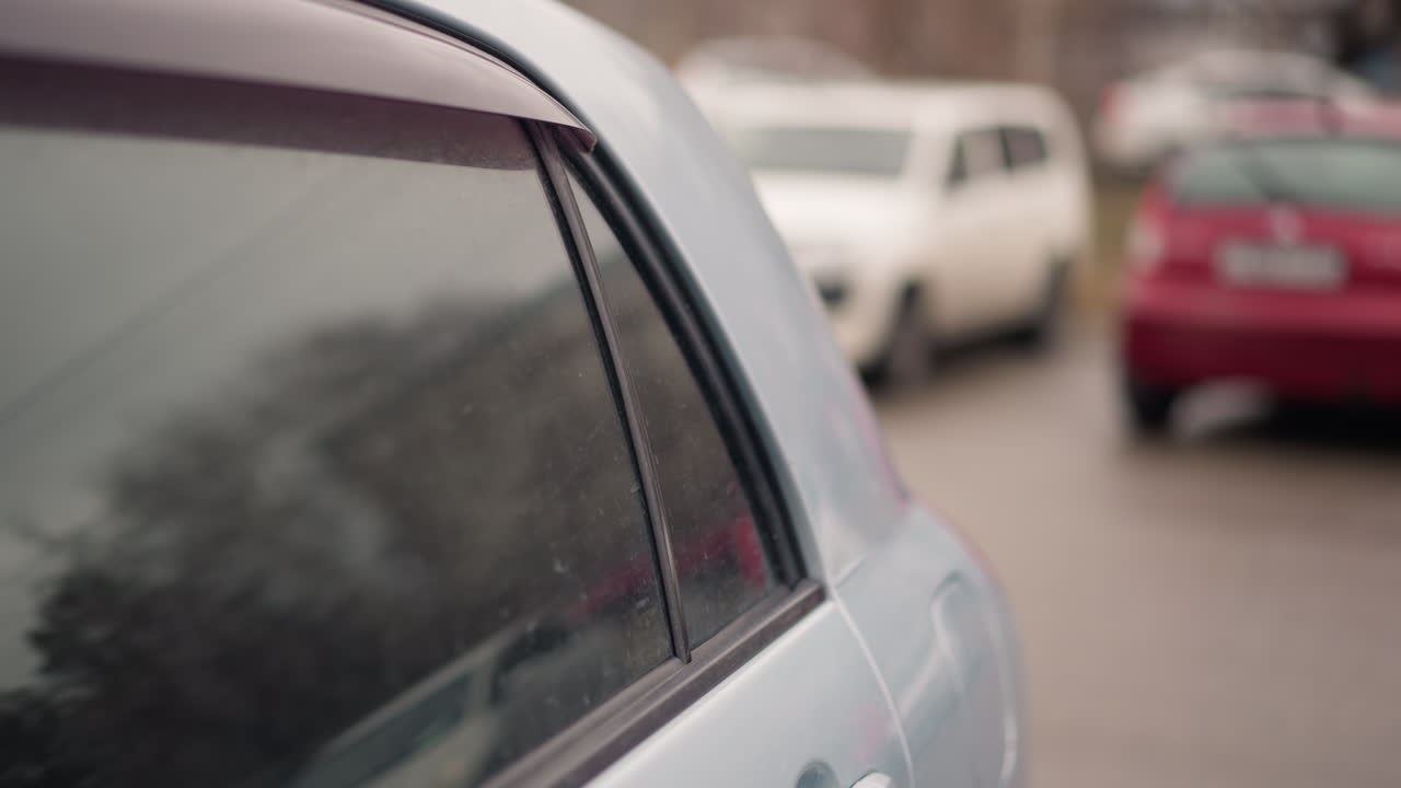 una vista cercana de coches estacionados con otro coche pasando en el fondo, el enfoque está en la ventana lateral de un coche de ceniza