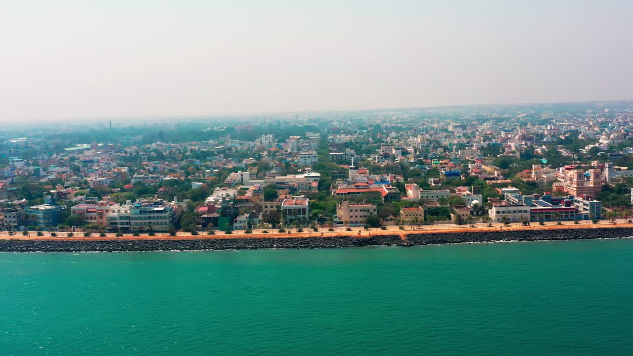 La Goubert Beach Road di Puducherry costeggia la Baia del Bengala turchese con un denso sviluppo urbano visibile nell'entroterra. Una lenta panoramica laterale a destra rivela il lungomare costiero e i quartieri residenziali.