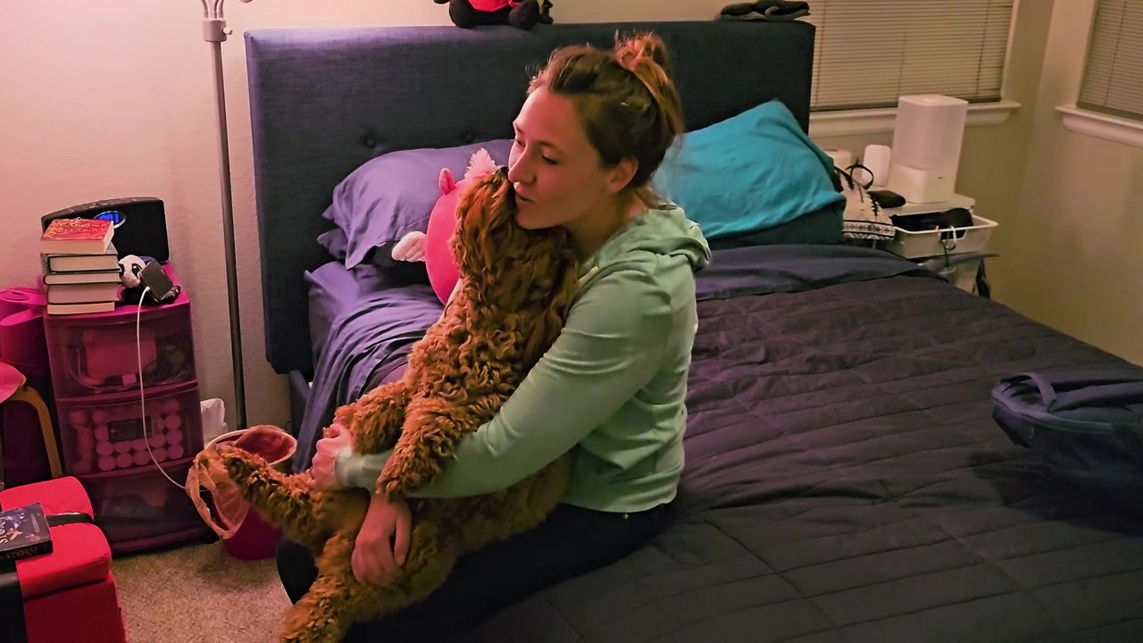 Woman dancing with her goldendoodle on the bed in a cozy bedroom, holding and hugging a curly brown dog, playful indoor lifestyle moment, pet companionship, love, and happy home scene.
