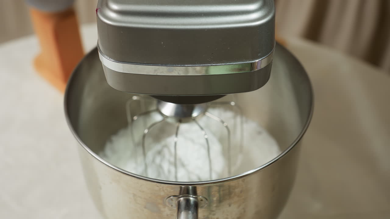 Closeup of electric mixer with white dough inside stainless bowl placed on kitchen table, whisk attachment clearly visible, other cooking tools blurred slightly in background