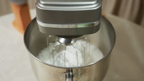 closeup of electric mixer with white dough inside stainless bowl placed on kitchen table, whisk attachment clearly visible, other cooking tools blurred slightly in background
