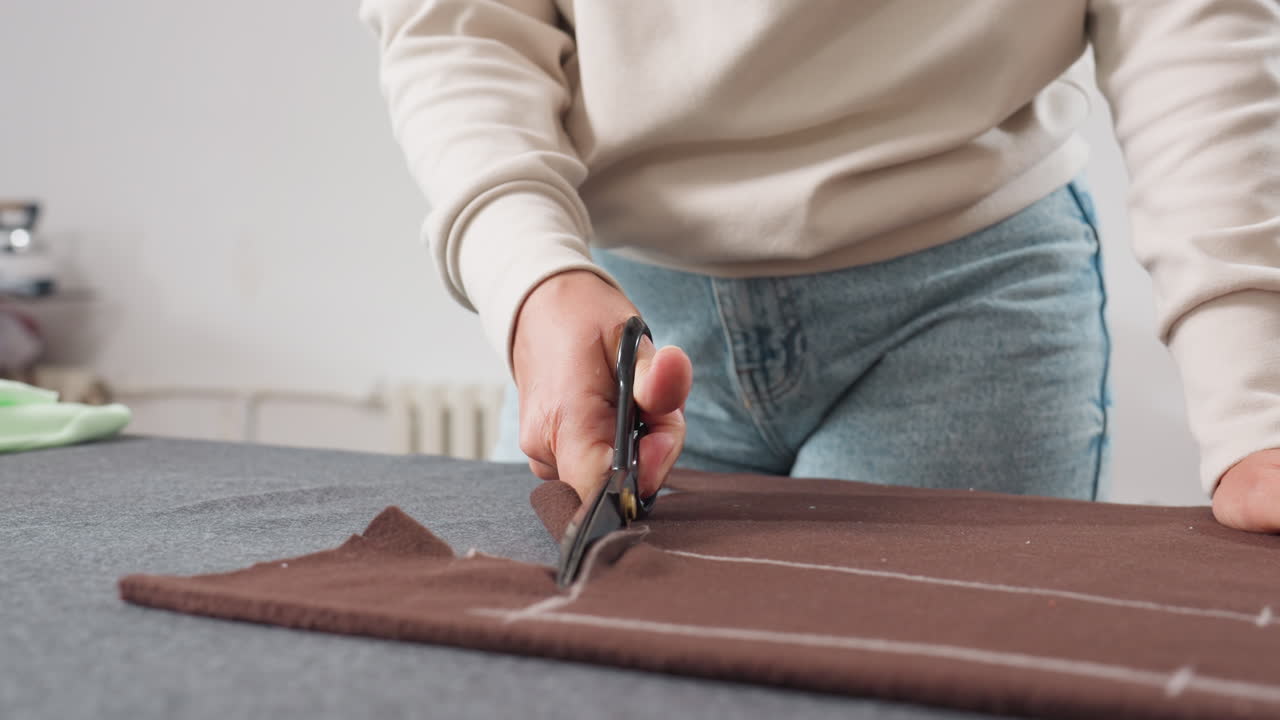Dress maker cutting brown cloth with large scissors following chalk outline on worktable, wearing light sweater and blue jeans, working carefully to create accurate fabric pieces in sewing studio