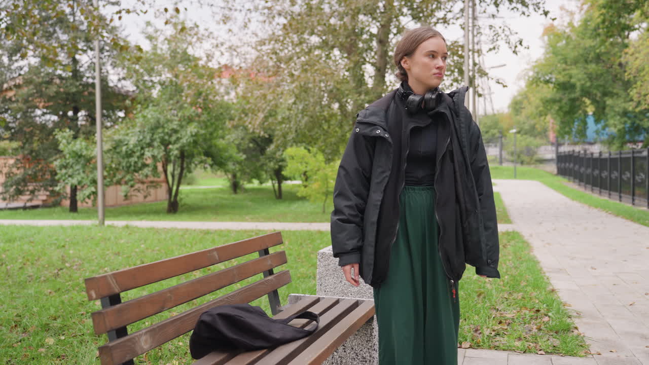 Giovane donna bianca che cammina su un sentiero verso una panchina in un parco urbano in un pomeriggio autunnale nuvoloso, indossa una giacca scura e cuffie, con uno zaino in spalla. Si ferma mentre un pendolare appoggia lo zaino e si avvicina