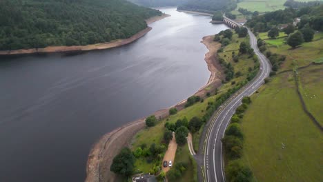 Drone footage glides above Ladybower Reservoir, revealing a winding road, lush green fields, and calm water under soft, overcast morning light