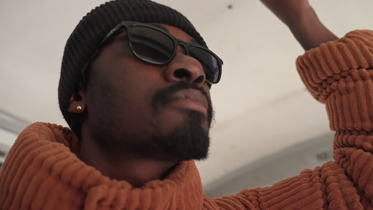 Black man on bus holding strap, wearing knit beanie and dark sunglasses, chunky rust sweater, backpack strap visible, candid closeup sequence showing introspective gaze and slight smile, morning city