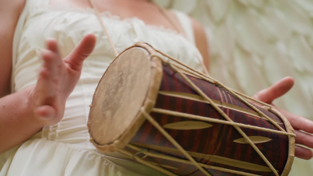 Close up of goddess with soft white wings gently beating talking drum in lush forest, hands moving gracefully with rhythm under warm daylight, expressing divine harmony