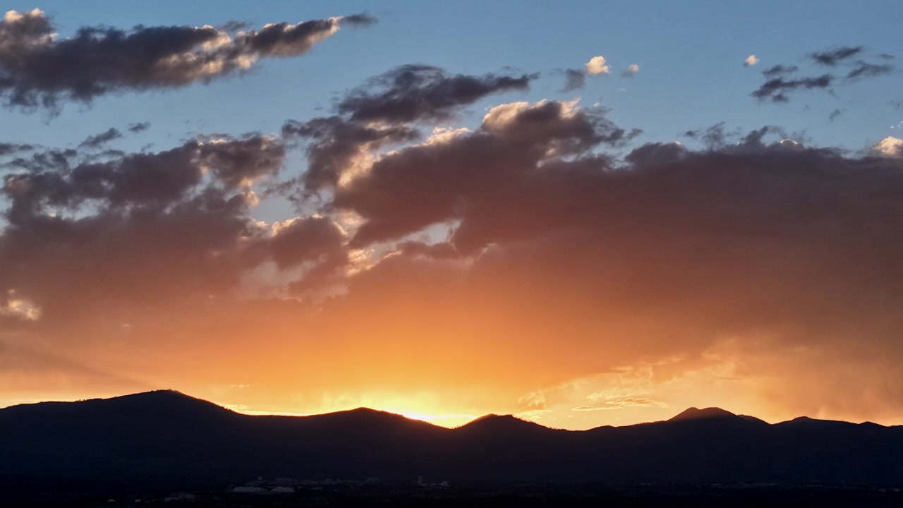 4K UHD drone timelapse capturing radiant golden orange dusk colors fading after sunset. A brilliant glowing light illuminates heavy evening clouds above a silhouetted Southwestern mountain range.