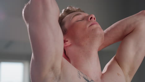 shirtless confident teenager with eyes closed poses with hands behind head, revealing strong arms and chest tattoo while standing in softly lit room, showing calm and cool self-assurance and poise