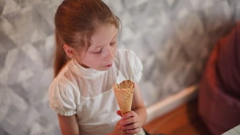 overhead view of young girl with long brown hair wearing white blouse happily holding ice cream cone, looking at it with joy before licking it, sitting indoors near keyboard with patterned background