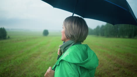 close-up of woman in green raincoat holding an open umbrella while looking into the distance, walking through an open green field under a cloudy sky, the background features vast grassy plains