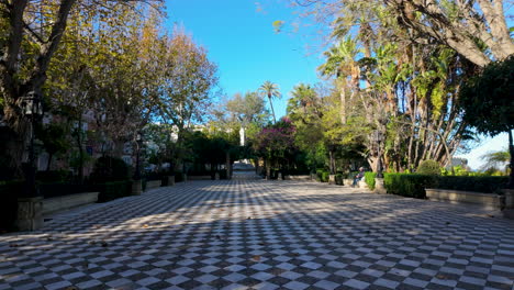 Ein-Breiter,-Von-Verschiedenen-Bäumen-Und-Palmen-Gesäumter-Weg-In-Einem-Ruhigen-öffentlichen-Park-Mit-Klarem,-Blauem-Himmel,-Der-Zu-Einem-Gemütlichen-Spaziergang-Oder-Einem-Friedlichen-Rückzugsort-In-Einer-Städtischen-Umgebung-Einlädt