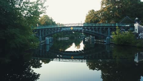 drone aerial moving forwards over river in a park during sunrise under a bridge with water reflections