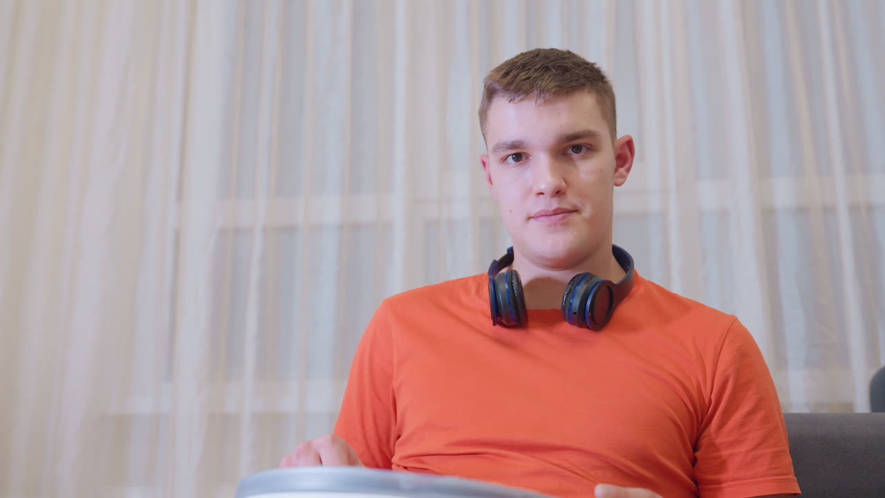 Jovem de camisa laranja com fones de ouvido no pescoço está sentado em um ambiente interno, trabalhando em um objeto com uma expressão calma e concentrada, tendo uma cortina como plano de fundo em um ambiente doméstico moderno.