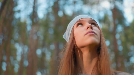 lady hiking through lush forest, looks around contemplatively, wearing blue bandana and backpack, surrounded by vibrant greenery and tall trees, with a soft, natural glow in background