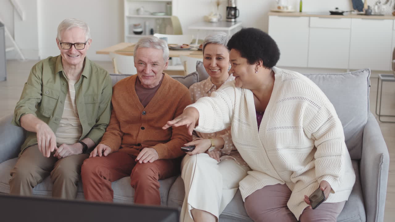 Medium long of four multiethnic diverse senior people sitting together on couch in living room, watching funny program on TV, talking and laughing