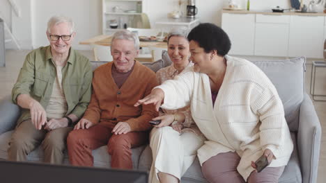 medium long of four multiethnic diverse senior people sitting together on couch in living room, watching funny program on tv, talking and laughing