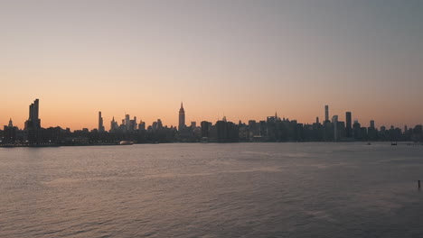 Antena:-Deslizándose-Sobre-El-Río-Este-Con-Vistas-Al-Horizonte-De-La-Ciudad-De-Manhattan-Nueva-York-En-La-Hermosa-Luz-Naranja-De-La-Puesta-Del-Sol-Del-Amanecer