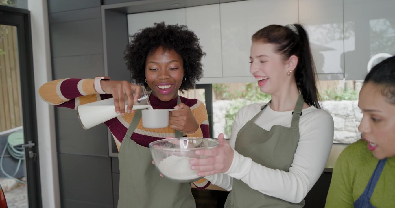 Donne diverse con grembiuli verdi che prendono e versano latte in una ciotola per cucinare sull'isola della cucina