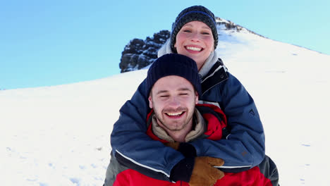 Lächelnder-Mann-Gibt-Seiner-Freundin-Ein-Huckepack-Im-Schnee