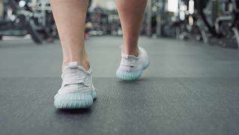 leg view of guy walking past gym equipment in health club, white sneakers stepping over rubber floor while blurred people exercise in background, conveying active lifestyle movement