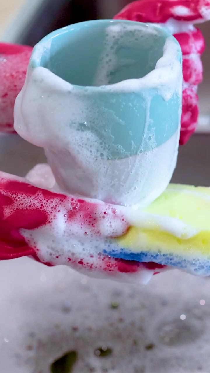 Close-up of hands wearing pink rubber gloves scrubbing a blue ceramic cup with soapy sponge.