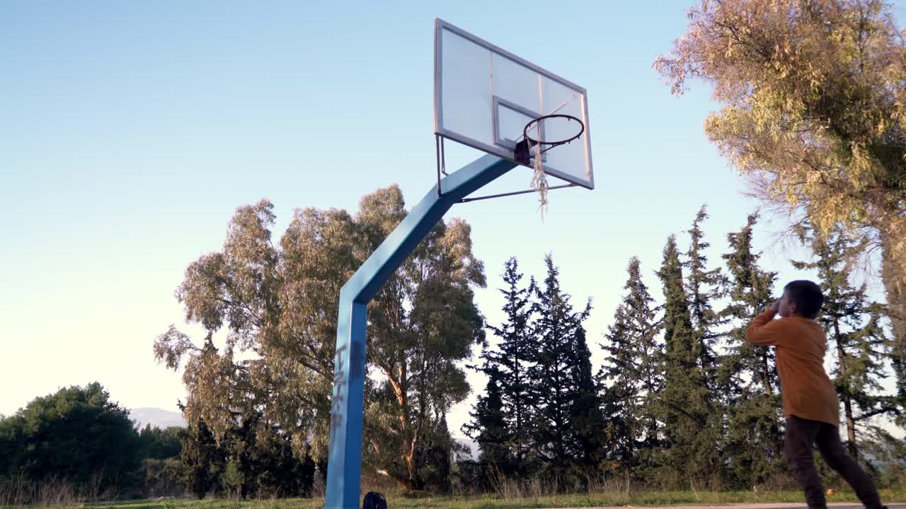 niño caucásico regateando y disparando una pelota de baloncesto al aire libre en un aro de cesta de la calle cerca de un bosque en parnitha, atenas, grecia