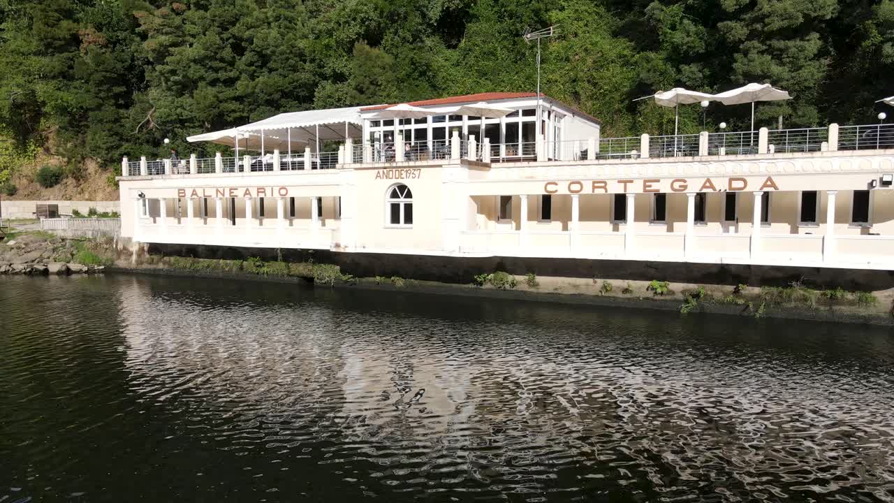 A serene riverside scene featuring a modern health resort nestled along the water’s edge. The building, painted in light tones, features an outdoor terrace with umbrellas and seating areas
