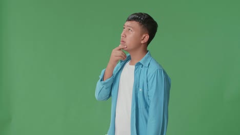 Side View Of Young Asian Boy Thinking About Something And Looking Around Then Raising His Index Finger And Eyes Wide Open While Standing On Green Screen In The Studio