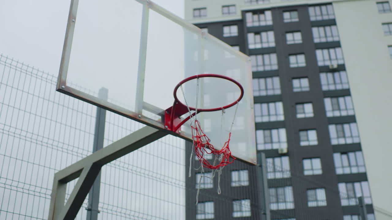 Canestro da basket vuoto su caseggiati, rete rossa logora e tabellone metallico incorniciato da una recinzione a maglie, cielo nuvoloso e pavimentazione in cemento che evocano quieta solitudine e un quartiere grintoso