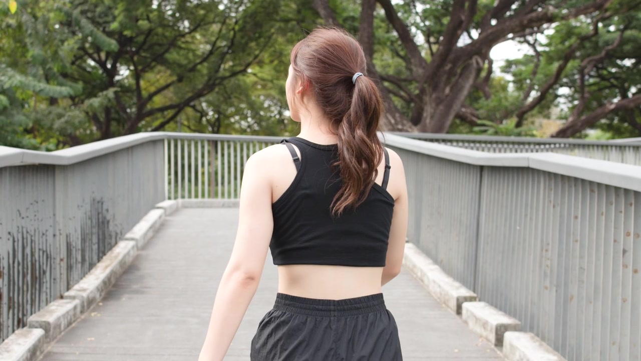 Rear view of a woman jogging across a modern bridge surrounded by lush green trees