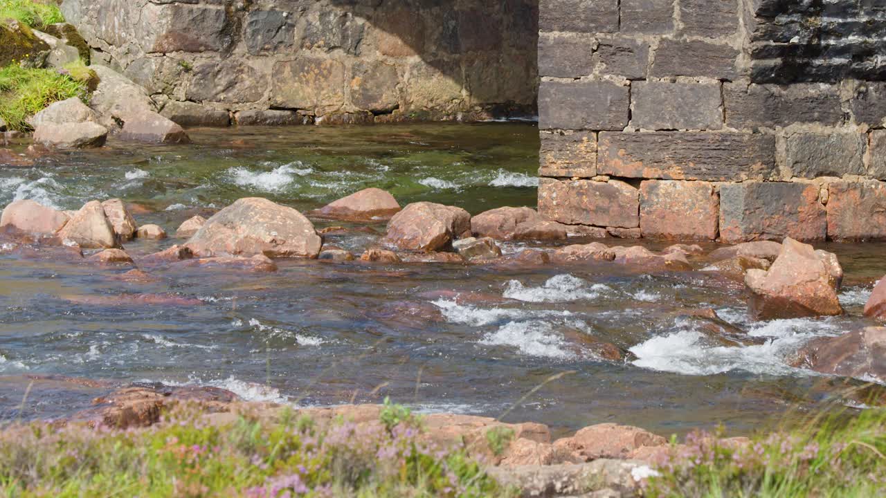 Camera slowly pans across historic stone bridge arch above flowing river, bright natural daylight